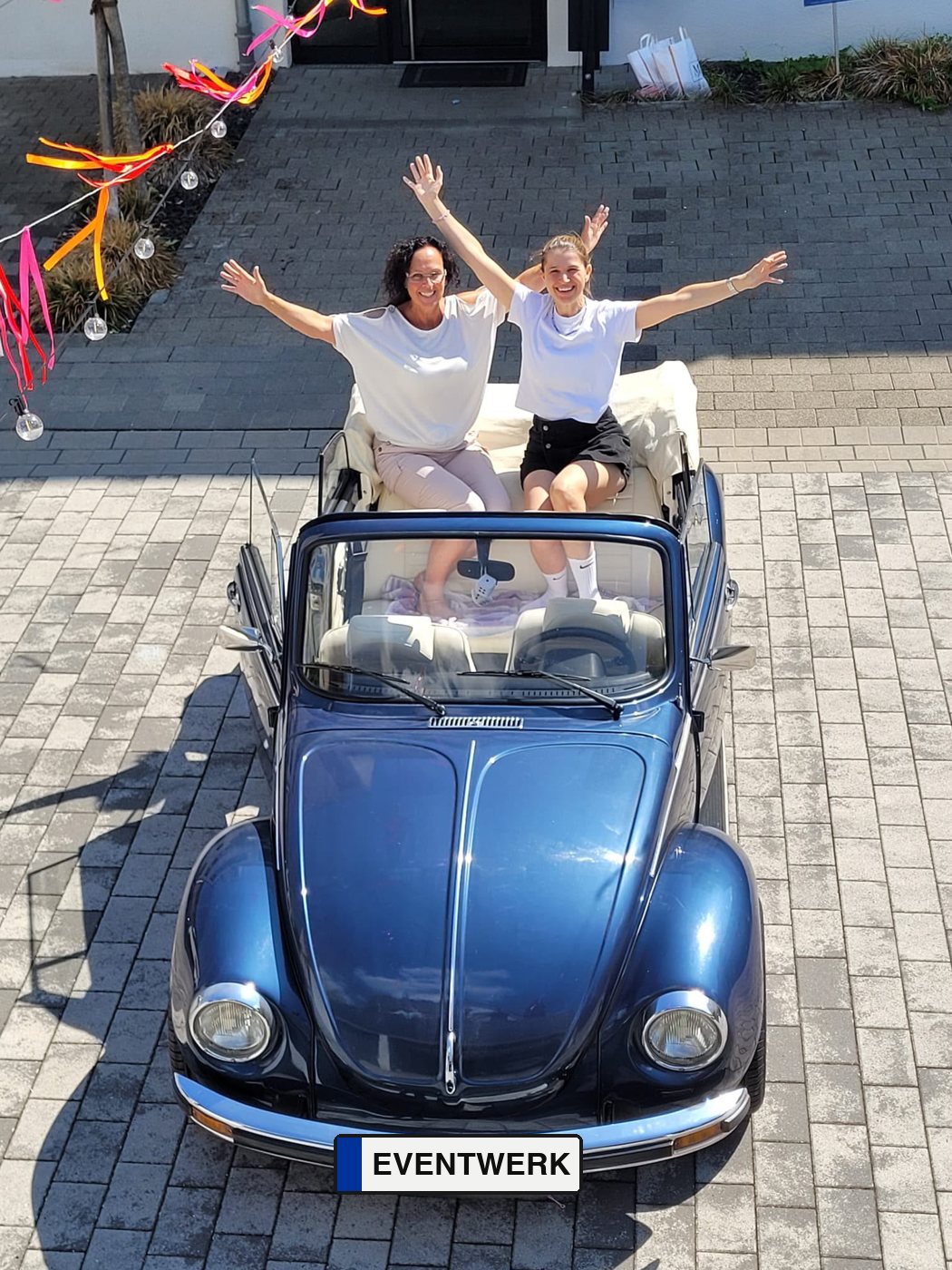Birgit und Carina jubeln mit ausgestreckten Armen in ihrem blauen Käfer